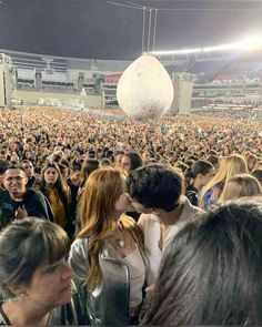 two people kissing each other in front of a large crowd at a concert with an enormous white ball hanging from the ceiling