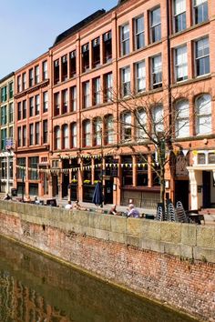 people sitting on benches along the side of a body of water in front of brick buildings