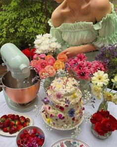 a woman sitting in front of a table filled with cakes and flowers next to bowls of strawberries