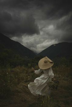 a woman in a white dress and straw hat walking through a field with mountains in the background