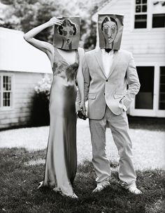 a man and woman standing next to each other in front of a house wearing paper bags on their heads