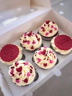 six cupcakes with white frosting and red hearts in a box on a table