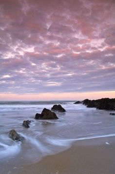 some rocks in the sand and water under a purple sky with clouds at sunset or dawn