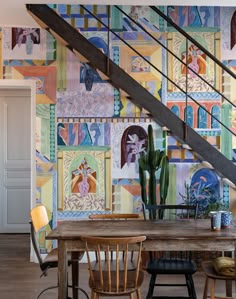 a dining room table with chairs under a stair case in front of a colorful wall