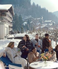 a group of people sitting around a table with drinks in front of some snow covered buildings