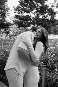 a man and woman standing next to each other in front of bushes with trees behind them