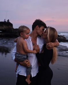 a man and woman kissing while holding a small child on the beach at sunset with people in the background