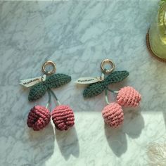 three crocheted cherries hanging from hooks on a marble counter top next to a vase