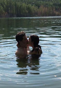 two people are kissing in the water near some trees and bushes, while one person has his face close to the other's head