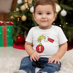 a baby sitting in front of a christmas tree wearing a t - shirt with the number one on it