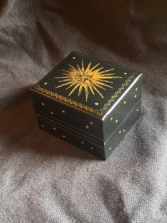 a small black box with gold designs on the lid sitting on a gray cloth covered surface