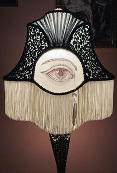 a lamp with an eye on it and fringes hanging from the bottom, in front of a wall