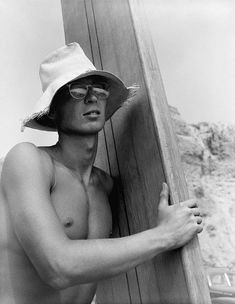 a shirtless man wearing a hat and glasses leaning against a wooden wall with his arms crossed