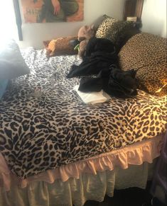 an unmade bed with leopard print sheets and clothes on it, in front of a painting