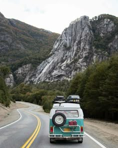 an old vw bus driving down the road in front of some mountains and trees