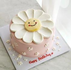 a pink and white birthday cake with a smiley face on the top is decorated with flowers