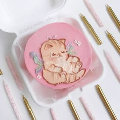 there is a pink cake with a cat on it and some gold pens next to it