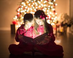 two young children sitting on the floor reading a book in front of a christmas tree