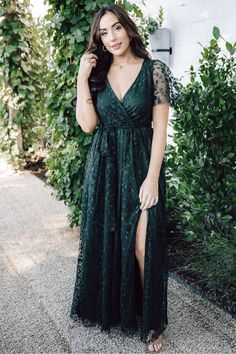 a woman wearing a green dress with sheer sleeves and thigh high slits, standing in front of greenery