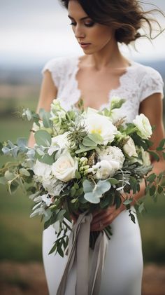 a woman holding a bouquet of white flowers and greenery in her hands while standing outside