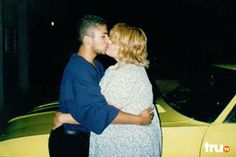 a man and woman are kissing in front of a yellow car at night with their arms around each other