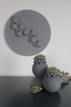 two gray vases with plants in them on a black table next to a white wall