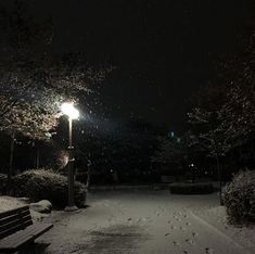 a bench sitting in the middle of a snow covered park at night with lights on