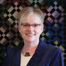 a woman wearing glasses and a blue jacket smiles at the camera while standing in front of a quilted wall