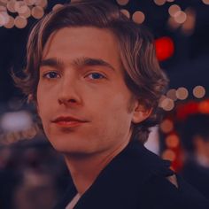 a man with long hair and blue eyes looks into the distance while standing in front of christmas lights