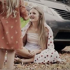 a woman sitting on the ground next to a car and talking to another woman in front of her