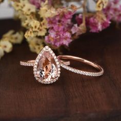 an image of two wedding rings on a table with flowers in the background and one ring has a pear shaped morganite surrounded by small diamonds