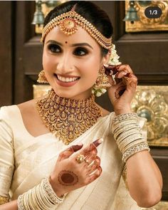 a woman in a white sari with gold jewelry on her neck and hands holding a flower