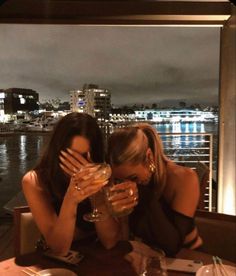 two women sitting at a table with wine glasses in front of their faces, looking out over the water