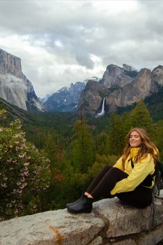 a woman sitting on top of a rock next to a forest filled with tall mountains