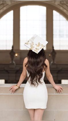 a woman in a white dress and graduation cap looking out over a wall with her hands on her hips