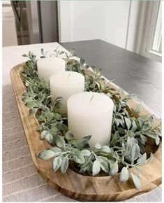 three white candles are sitting in a wooden bowl with greenery on the table top