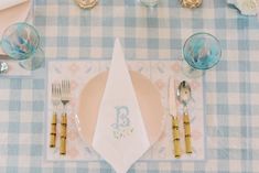 a place setting with blue and white checkered table cloth, gold rimmed utensils and napkins