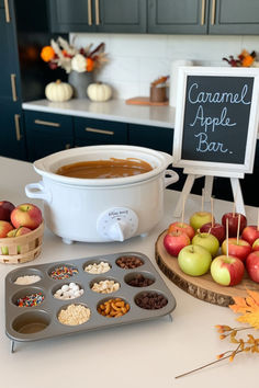 Cozy caramel apple bar setup perfect for a November baby shower, featuring a white slow cooker filled with warm caramel, a selection of apples on a wooden board, and an array of toppings in a muffin tin. The setup is ideal for fall-themed November baby shower ideas and November baby shower food options. Fall Baby Shower Boy Theme, November Baby Shower Food Ideas, November Baby Shower Ideas Decoration, Diy Fall Baby Shower Decorations, Baby Shower Ideas Fall Girl, November Baby Showers, Fall Shower Food, Fall Themed Party Decorations, Pumpkin Gender Reveal Food Ideas