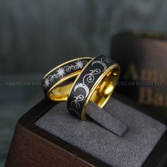 two gold wedding rings with black and white designs on them sitting on a wooden stand