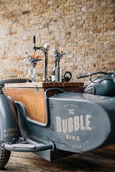 an old fashioned car is parked in front of a brick wall and table with wine glasses on it