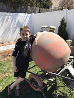 a young boy standing next to a large cement ball on a wheelbarrow in the yard