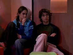 two people sitting on a couch in a room with pink walls and flooring, one person is looking at the camera