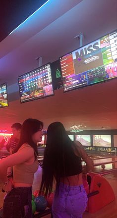 two girls are playing bowling in a bowling alley with neon lights on the ceiling and televisions above them