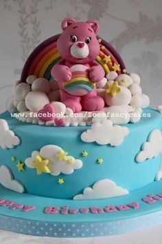 a blue cake with a pink teddy bear on top and rainbow in the sky above it