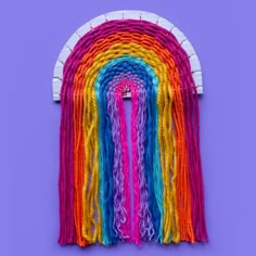 a rainbow wall hanging on a purple wall with the colors of yarn in different shades