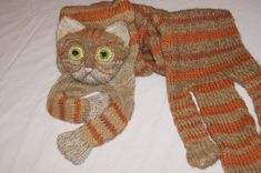 an orange and brown knitted cat laying on top of a white bed next to a scarf