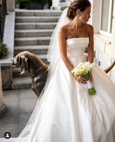 a woman in a white wedding dress standing next to a brown dog and holding a bouquet