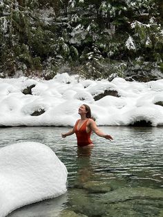 a woman in a red swimsuit wading through a river with snow on the bank