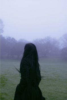 a woman with long black hair standing in a foggy field looking at the ground