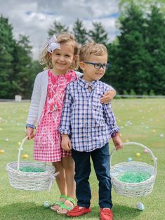 two young children standing next to each other holding baskets with colored eggs in them on the grass